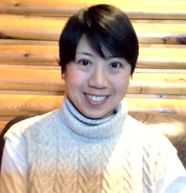 Photo of a woman from the shoulders up with short black hair and dark eyes, smiling at the camera. She is wearing a light colour high neck cable knit sweater against a wood grain background.